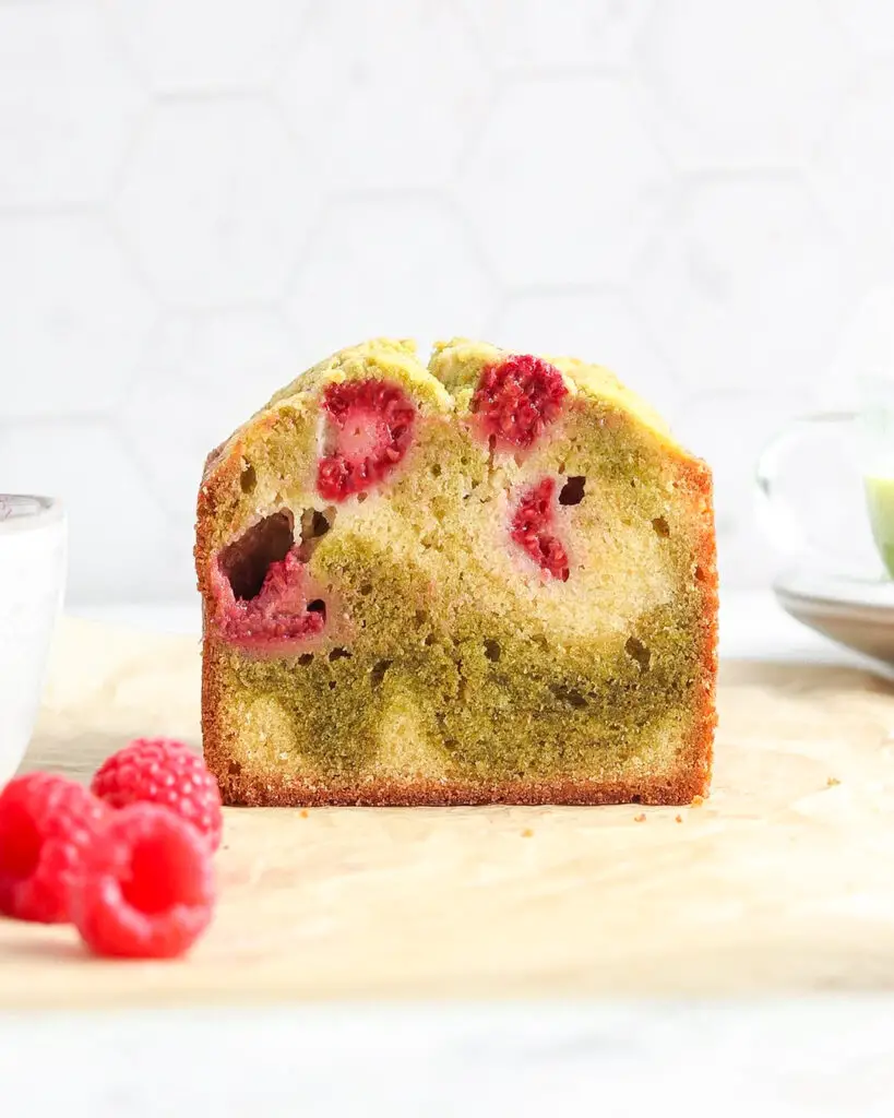 The cross section of a matcha raspberry swirl pound cake loaf, sitting on a piece of parchment paper surrounded by raspberries and a cup of matcha tea
