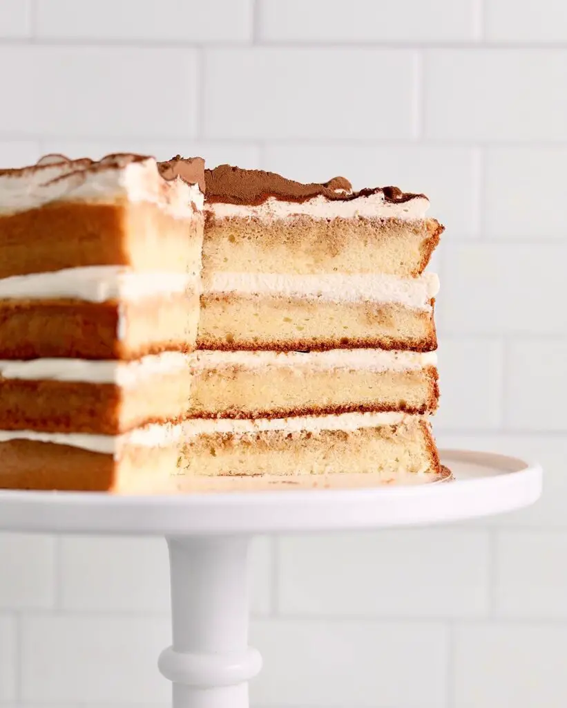 A tiramisu layer cake on a white cake stand with mascarpone whipped cream frosting, dusted with cocoa powder