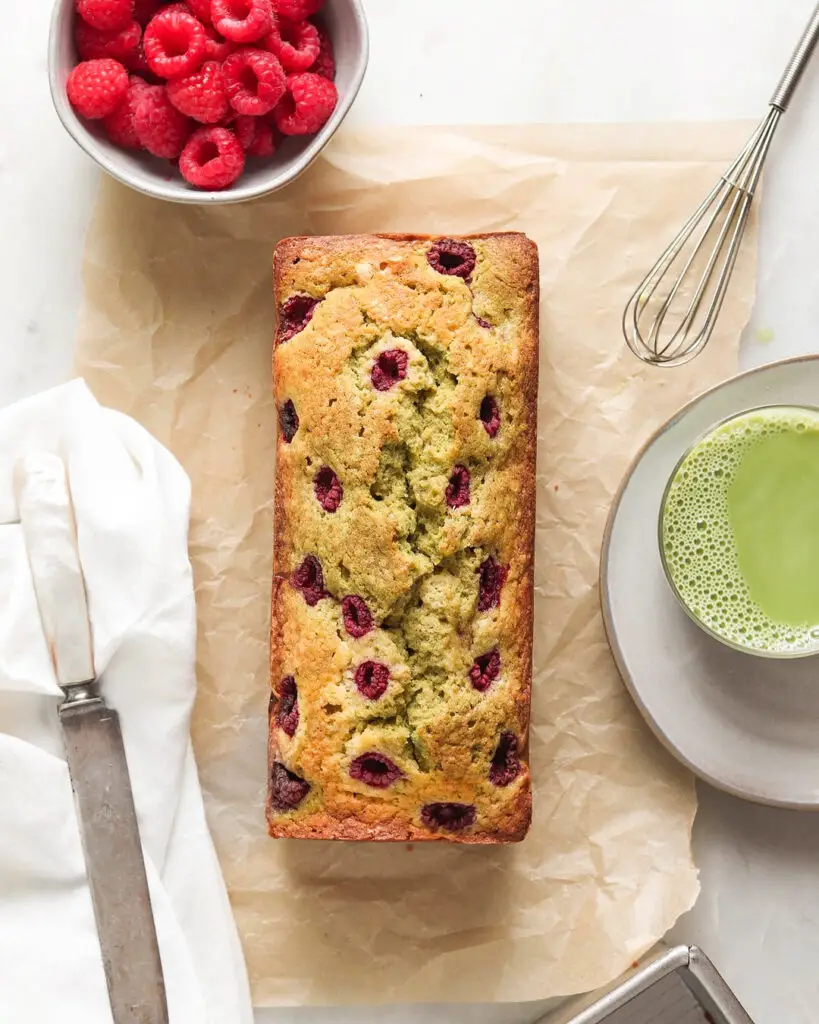 Overhead view of a matcha raspberry swirl pound cake loaf, sitting on a piece of parchment paper surrounded by raspberries and a cup of matcha tea