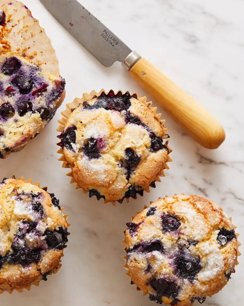 Four blueberry muffins scattered on a white countertop and a small knife with a wooden handle 