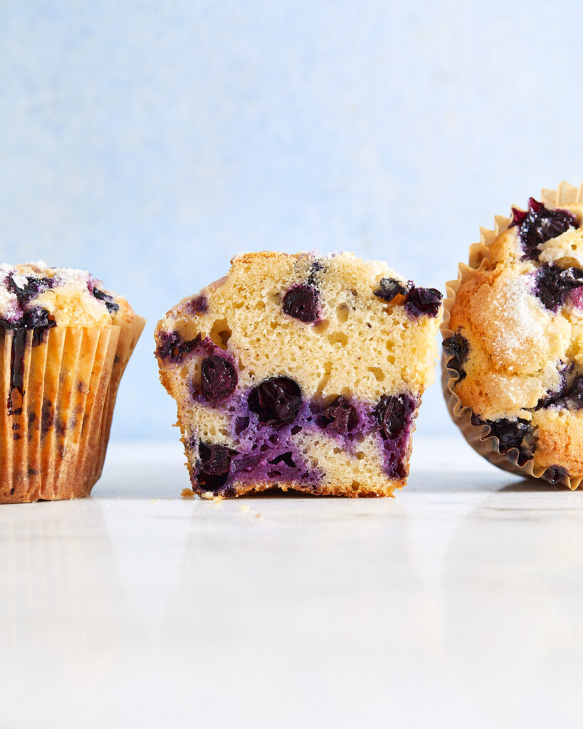 large bakery style muffins in a row, one upright with its liner on, one cut in half to reveal the inside, and one on its side showing the top