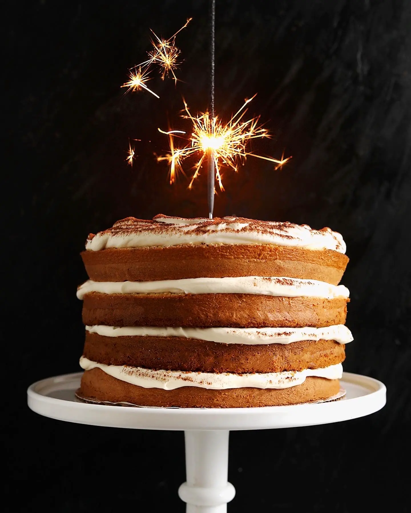 a cake on a white cake stand with layers of cake and mascarpone whipped cream. A burning sparkler is sticking out of the top