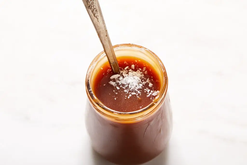 salted caramel sauce in a small jar with a spoon in it and a sprinkle of flakey sea salt