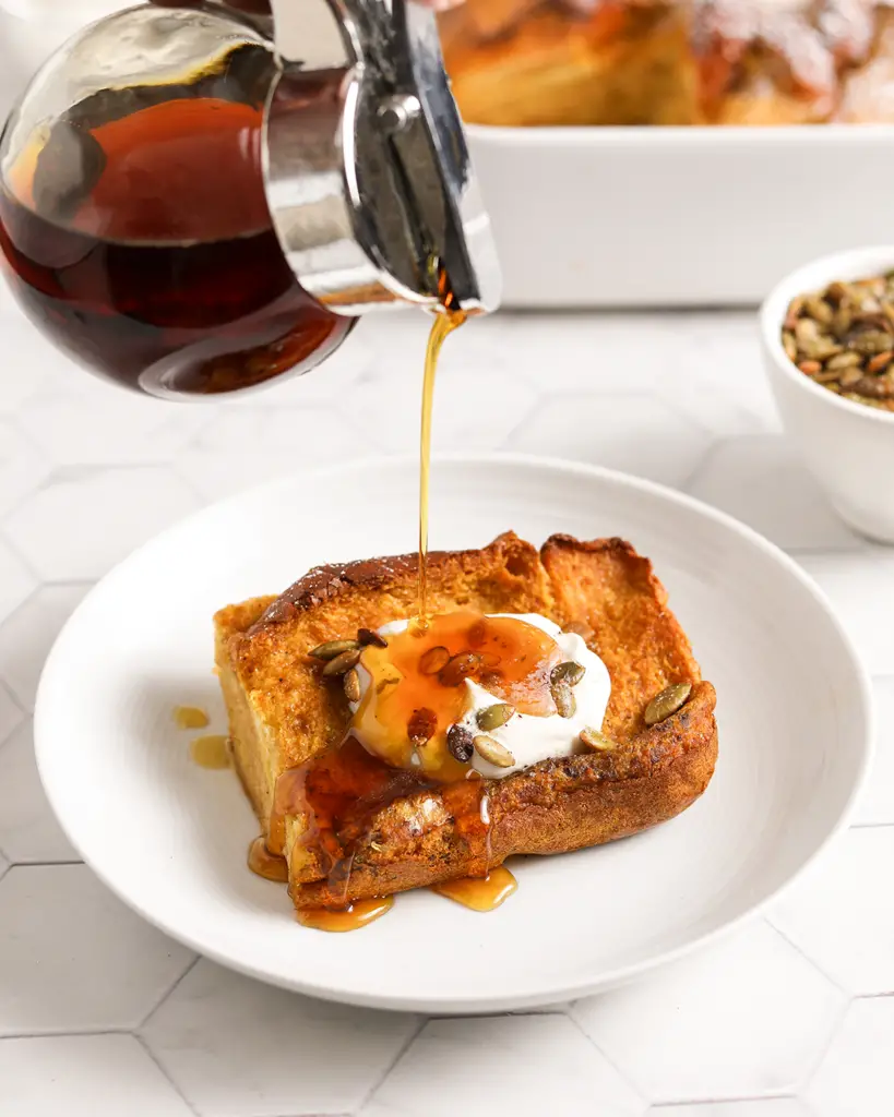 a slice of overnight pumpkin french toast bake on a plate, with maple syrup being drizzled over it, topped with spiced pumpkin seeds and maple whipped cream