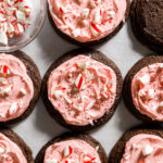 a dozen chocolate chocolate cookies frosted with pink peppermint buttercream sprinkled with crushed up candy canes
