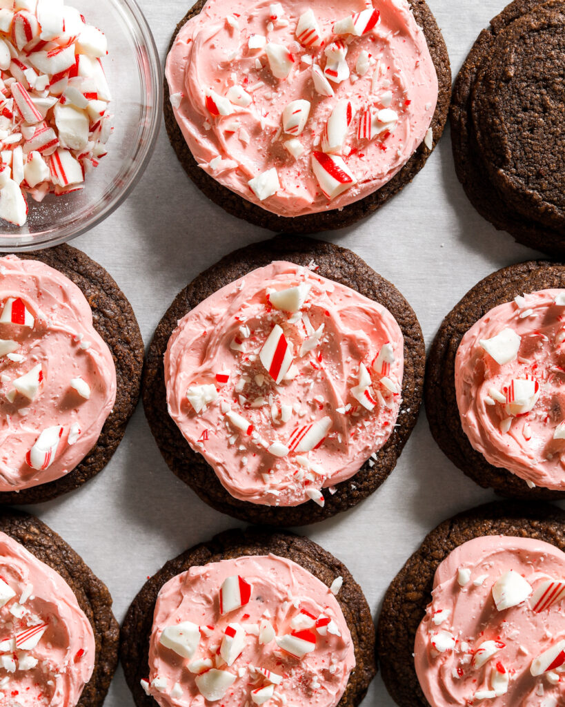 a dozen chocolate chocolate cookies frosted with pink peppermint buttercream sprinkled with crushed up candy canes