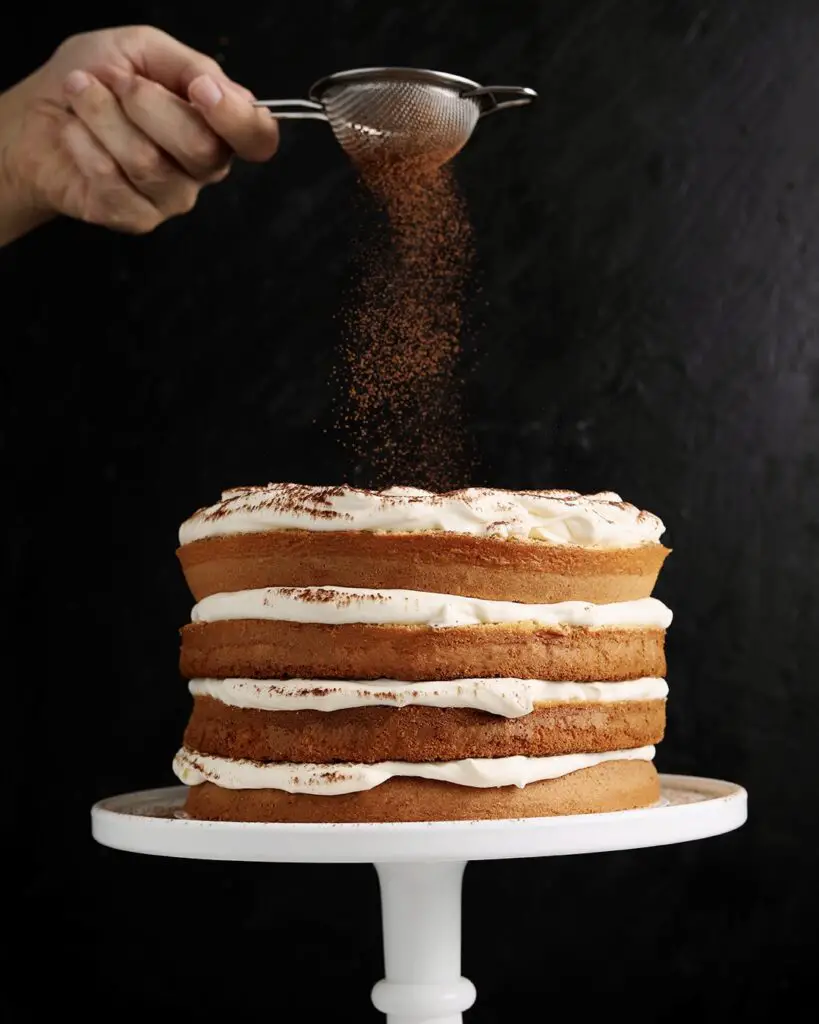 A tiramisu layer cake on a white cake stand with mascarpone whipped cream frosting. Someone is using a fine mesh sieve to dust cocoa powder on top