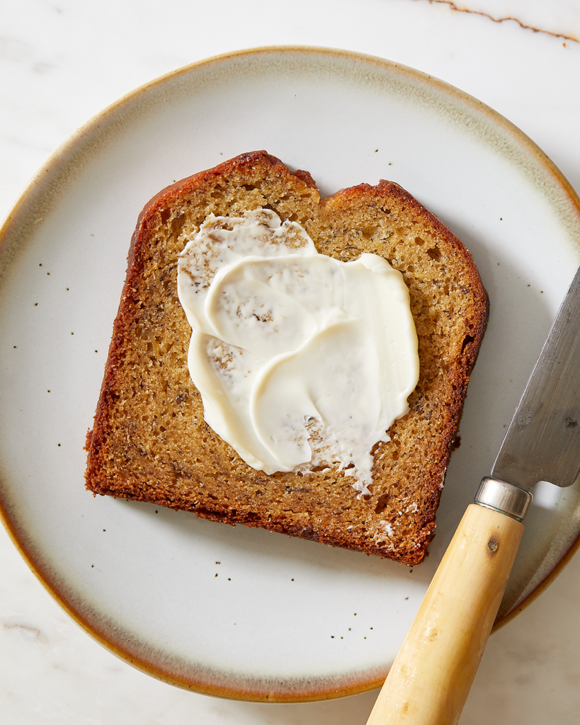 a slice of banana bread with a smear of butter sits on a blue dessert plate with a small knife next to it. 