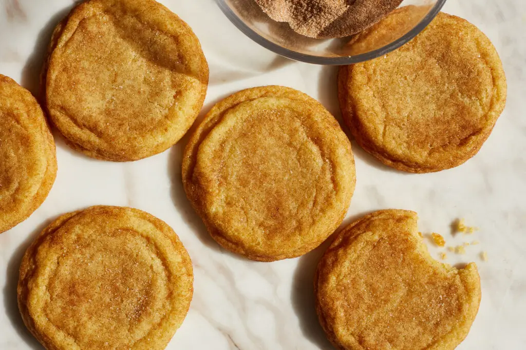 chewy snickerdoodle cookies on a white marble surface next to a bowl of cinnamon sugar