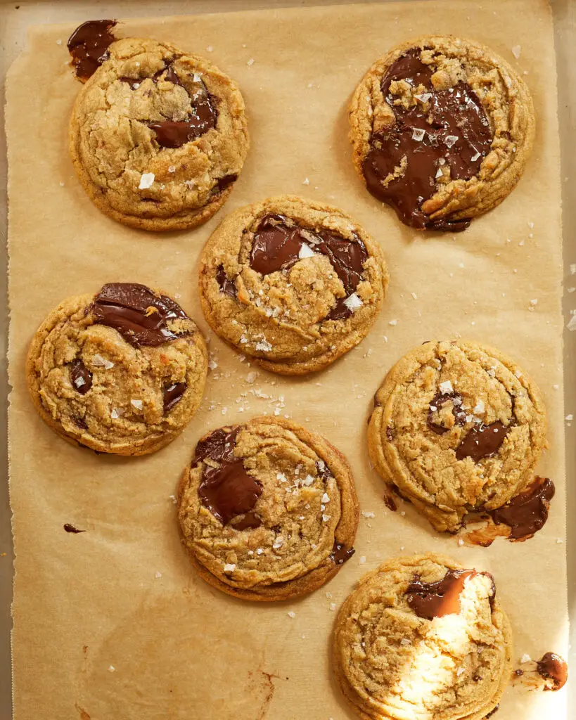 chocolate chip cookies with big puddles of chocolate and sprinkled with flakey salt on a baking sheet lined with tan parchment