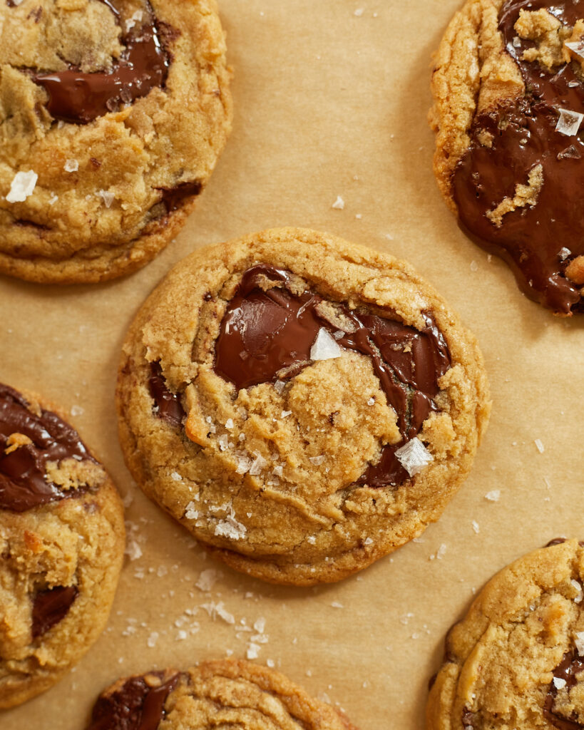 chocolate chip cookis with big puddles of chocolate and sprinkled with flakey salt on a baking sheet lined with tan parchment 