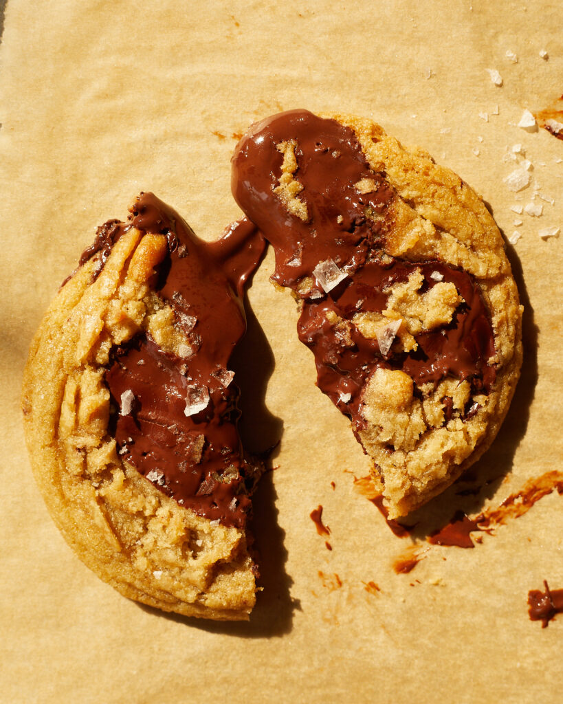 chocolate chip cookis with big puddles of chocolate and sprinkled with flakey salt on a baking sheet lined with tan parchment 