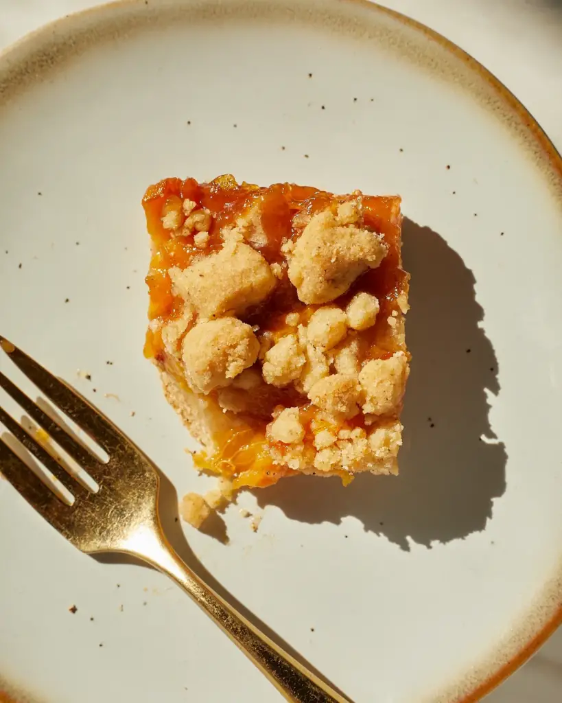 A square of peach crumble bar on a blue dessert plate next to a brass fork