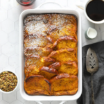 overnight pumpkin french toast bake casserole, dusted with powdered sugar, with a coffee cup, pumpkin seed topping and maple syrup on a countertop