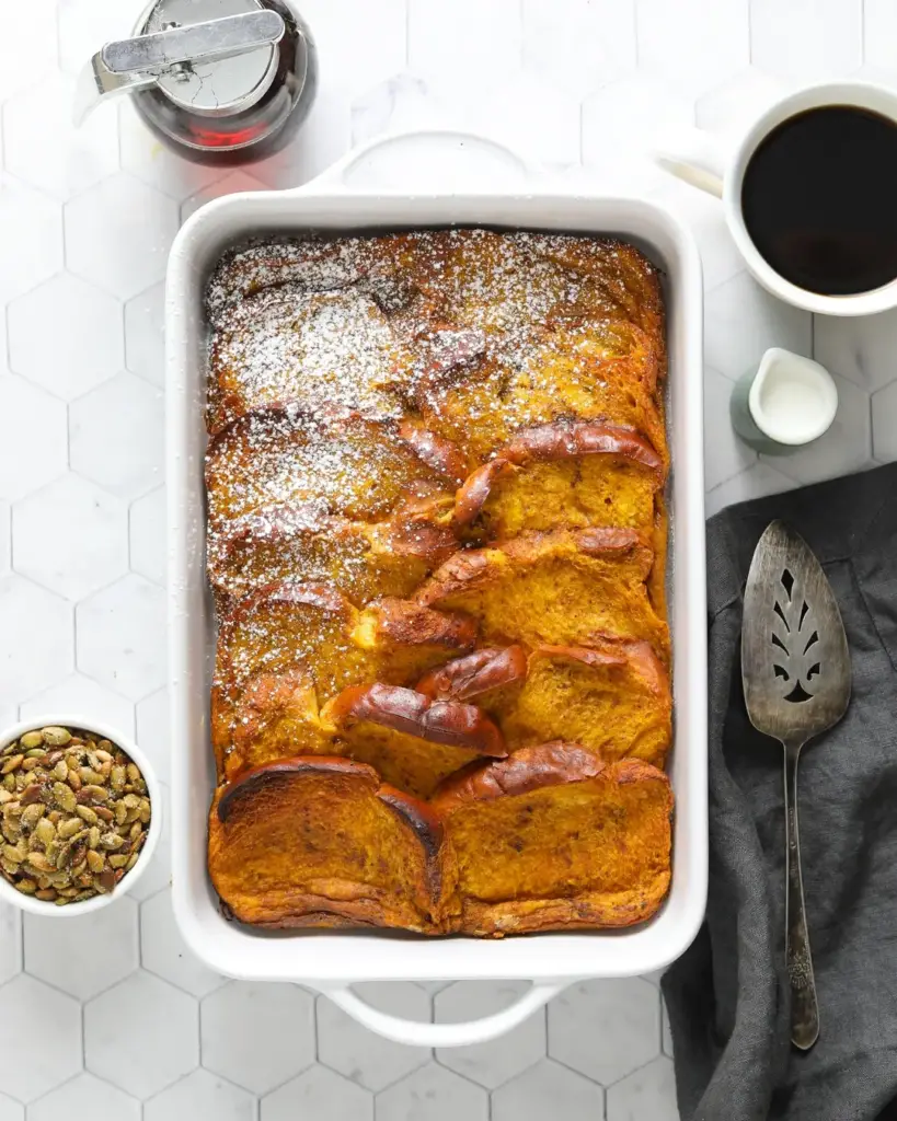 overnight pumpkin french toast bake casserole, dusted with powdered sugar, with a coffee cup, pumpkin seed topping and maple syrup on a countertop