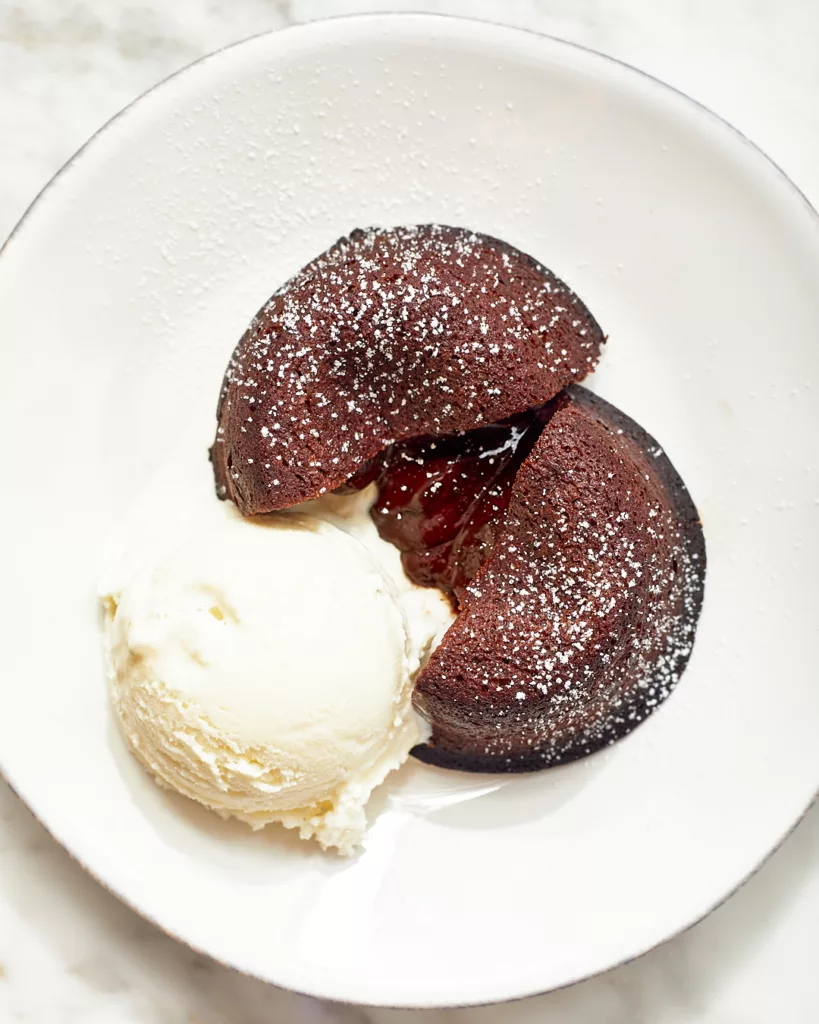 a mini chocolate lava cake cut in half with molten chocolate ganache oozing out of its center, next to a scoop of vanilla ice cream on a dessert plate