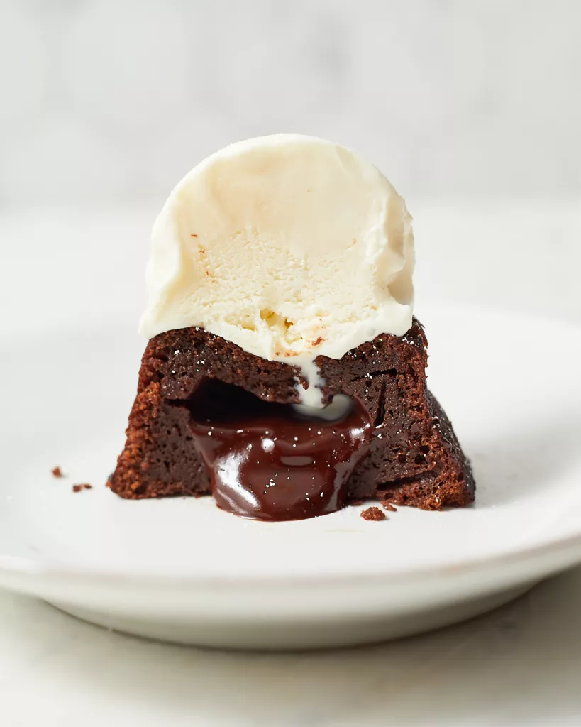 a mini chocolate lava cake cut in half with molten chocolate ganache oozing out of its center, sitting under a scoop of vanilla ice cream