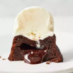 a mini chocolate lava cake cut in half with molten chocolate ganache oozing out of its center, sitting under a scoop of vanilla ice cream