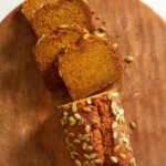 a loaf of pumpkin bread with pumpkin seeds or pepitas on top with slices cut off on a wooden serving tray