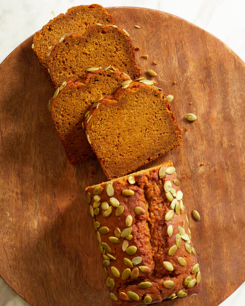 a loaf of pumpkin bread with pumpkin seeds or pepitas on top with slices cut off on a wooden serving tray 