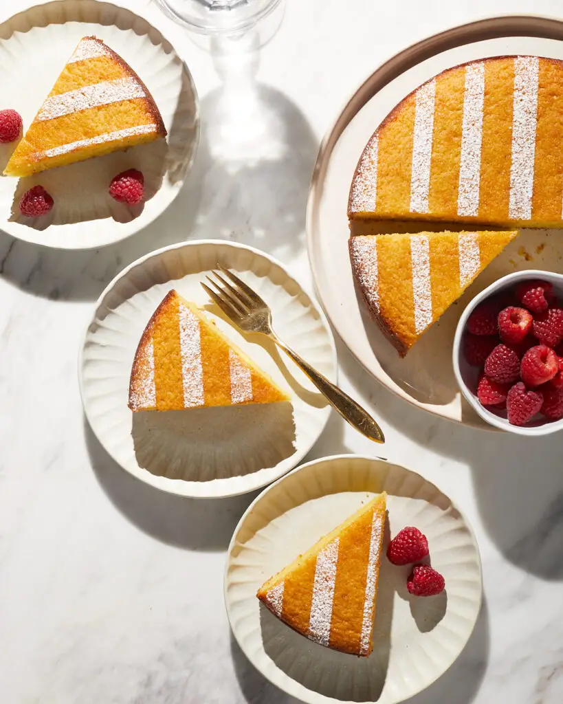slices of ricotta olive oil lemon snacking cake on white dessert plates with raspberries on a white marble countertop