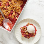 A scoop of vegan strawberry rhubarb crumble on a small white dessert plate with the full tin and a serving spoon next to it