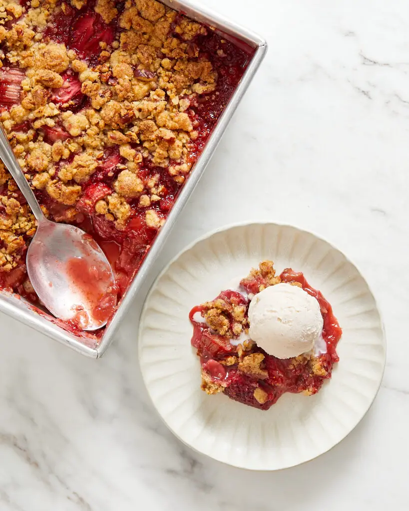 A scoop of vegan strawberry rhubarb crumble on a small white dessert plate with the full tin and a serving spoon next to it 