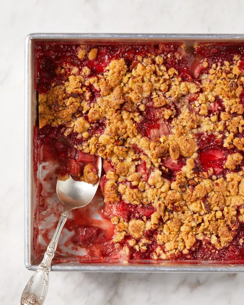 a 9x9 inch tin of vegan strawberry rhubarb crumble with a serving spoon dipping into it