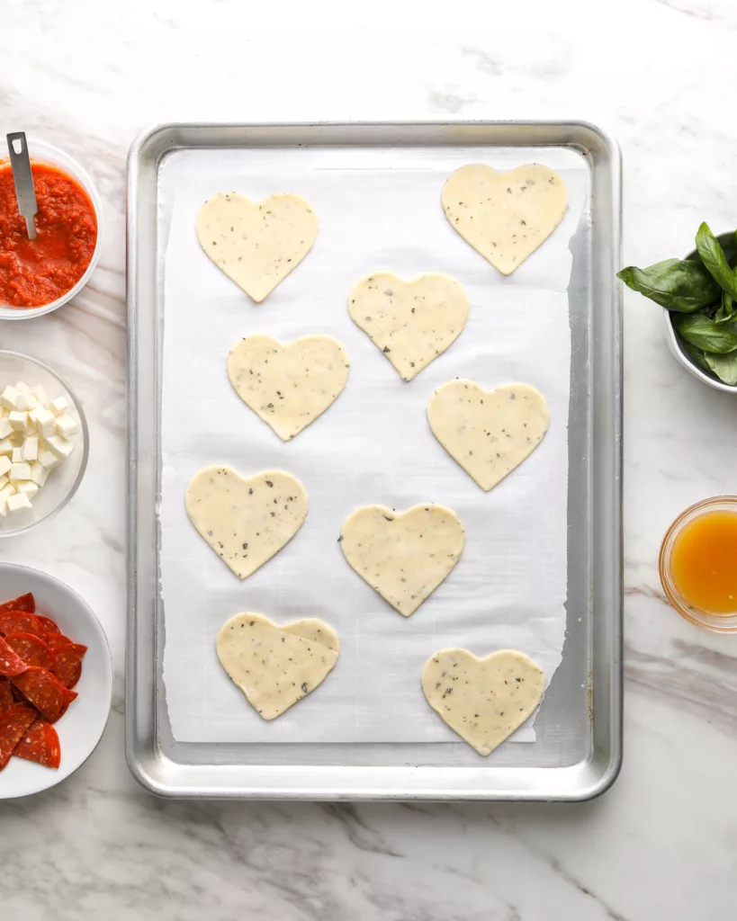 9 heart-shaped pie crusts sit on a baking tray surrounded by pizza ingredients
