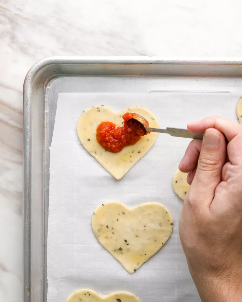 a hand dollops pizza sauce on heart shaped pie crust