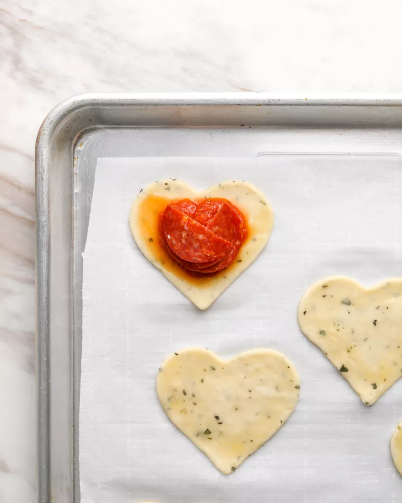 slices of pepperoni is placed on top of pizza sauce on heart shaped pie crust