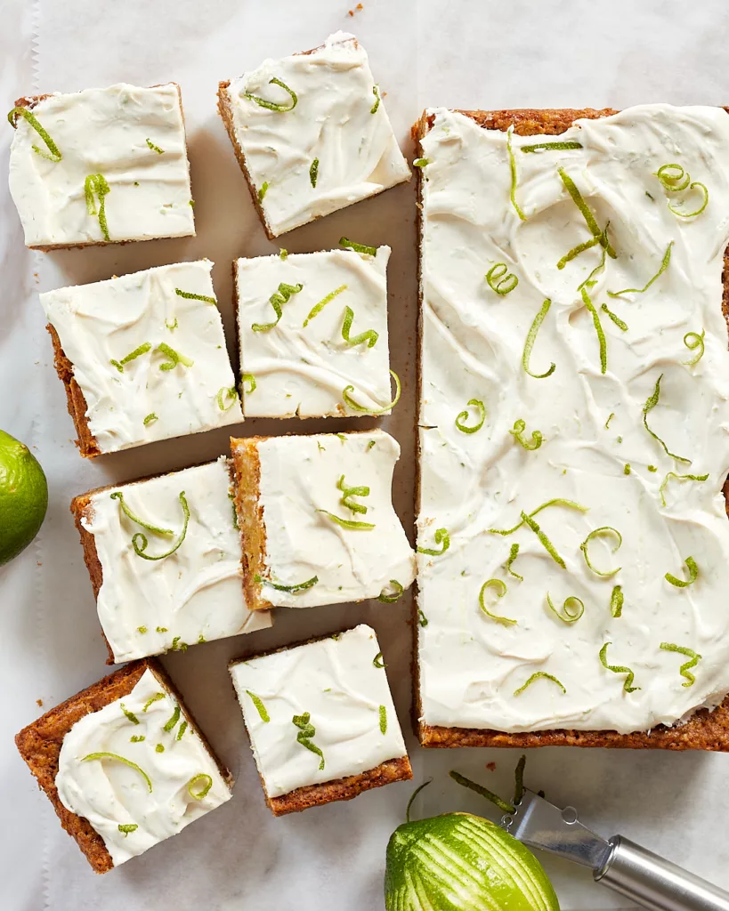 a tray of lime coconut bars with swirls of lime cream cheese frosting and curls of lime zest on top cut into slices on a white piece of parchment