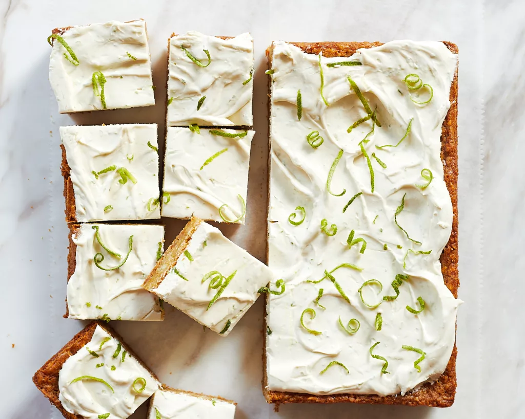 a tray of lime coconut bars with swirls of lime cream cheese frosting and curls of lime zest on top cut into slices on a white piece of parchment