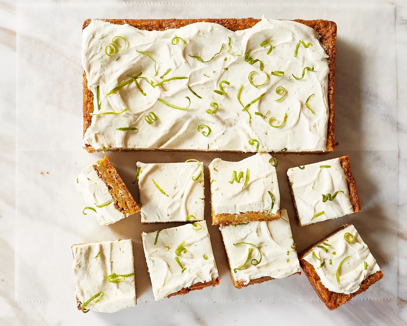 a tray of lime coconut bars with swirls of lime cream cheese frosting and curls of lime zest on top cut into slices on a white piece of parchment