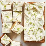 a tray of lime coconut bars with swirls of lime cream cheese frosting and curls of lime zest on top cut into slices on a white piece of parchment.