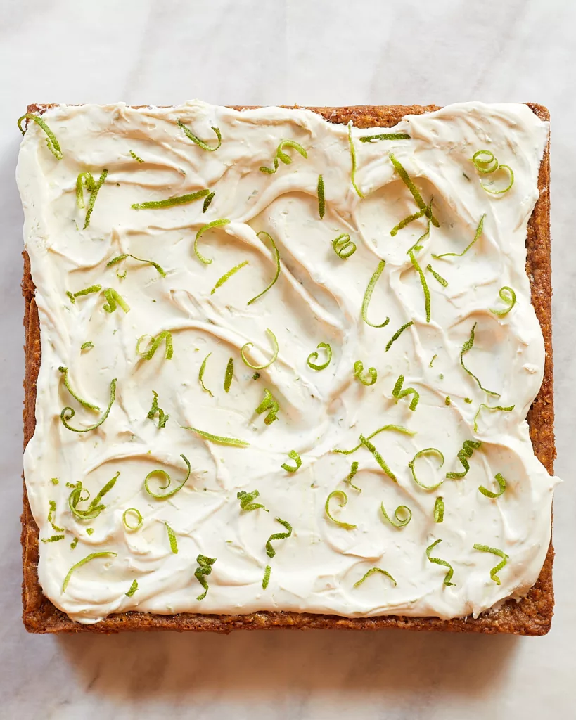 a 9 by 9 tray of lime coconut bars with swirls of lime cream cheese frosting and curls of lime zest on a white piece of parchment