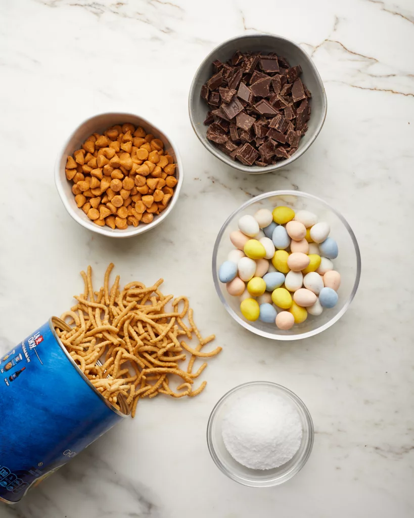 the ingredients for birds nest cookies like chocolate, butterscotch chips, chow mein noodles, salt, and chocolate candy eggs, on a white surface