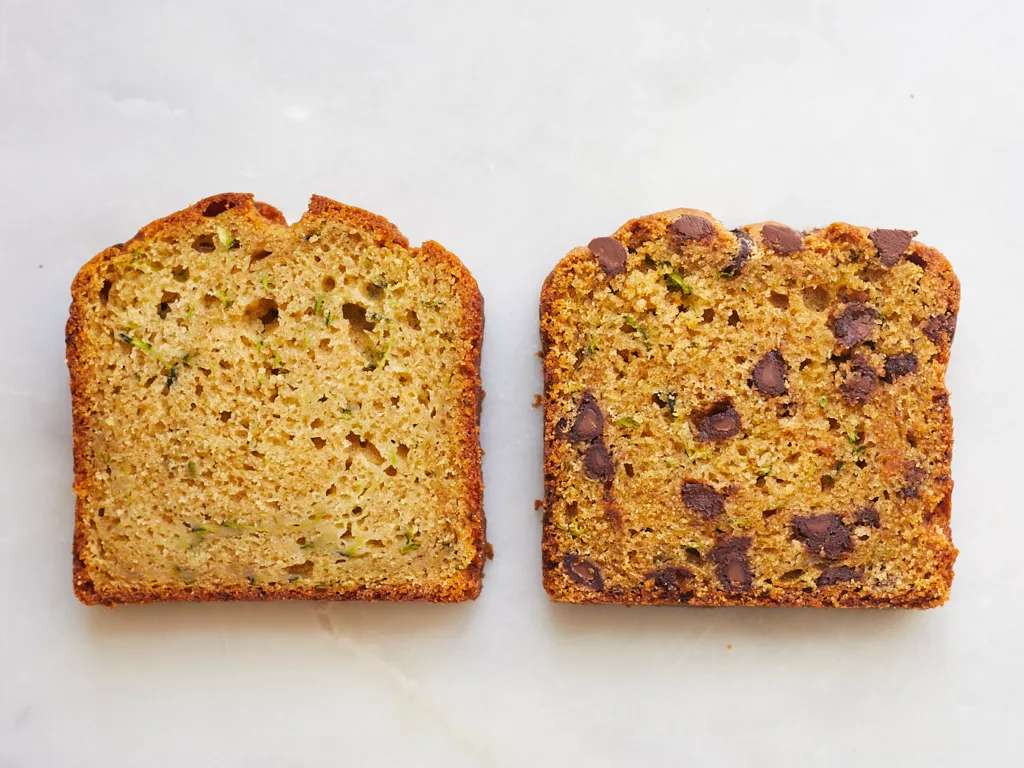 two slices of olive oill zucchini bread, one plain and one with chocolate chips sit on a marble background