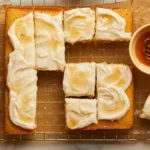 cornbread snacking cake cut into slices on a wire rack surrounded by honey and a spatula with cream cheese frosting on it