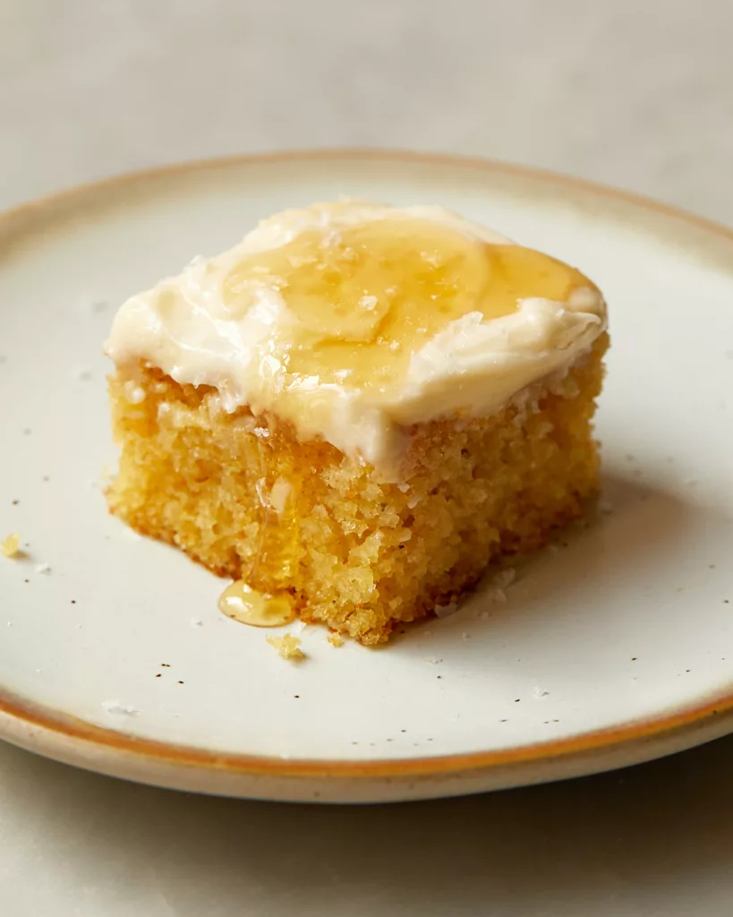 A slice of cornbread snacking cake with honey-cream cheesee frosting sitting on a dessert plate with a drizzle of honey running down the side