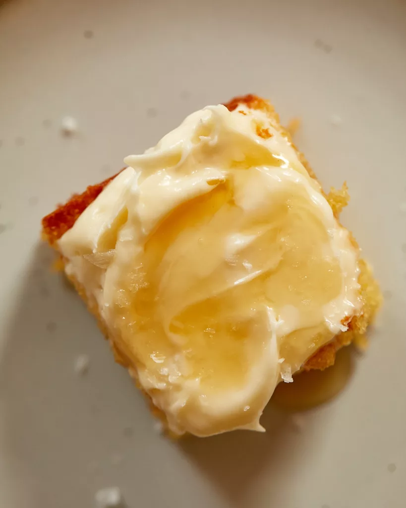 A slice of cornbread snacking cake with honey-cream cheesee frosting sitting on a dessert plate with a drizzle of honey running down the side