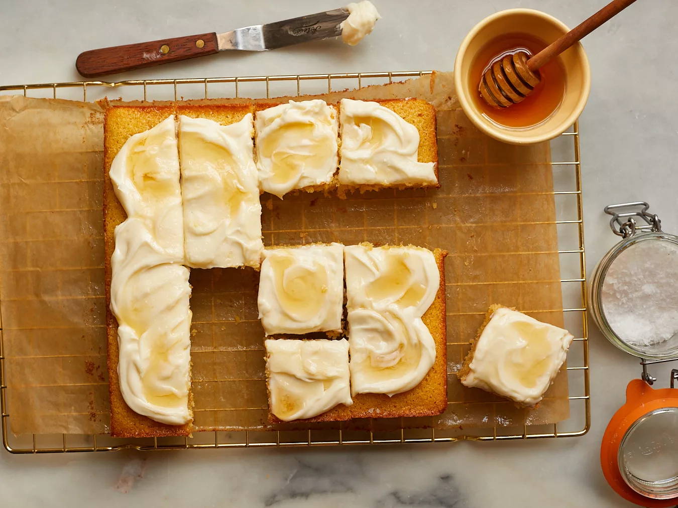 cornbread snacking cake cut into slices on a wire rack surrounded by honey, a jar of flakey salt, and a spatula with cream cheese frosting on it