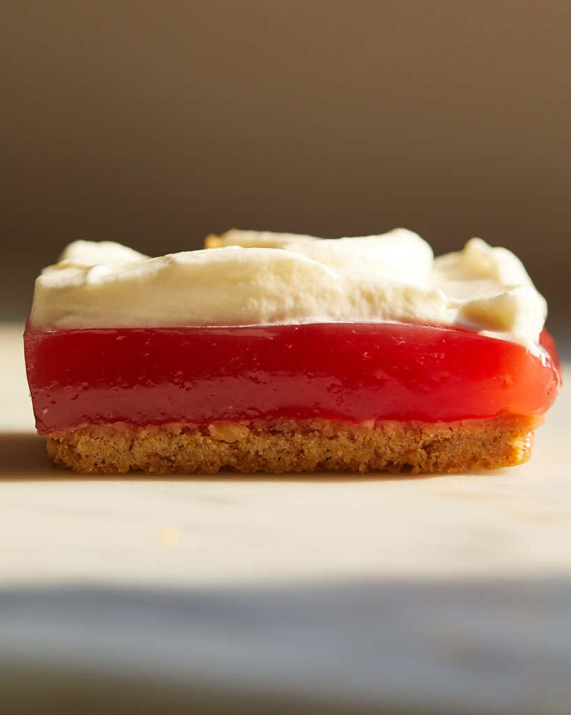 A watermelon strawberry jello bar with layers of pretzel shortbread and whipped cream sits on a white marble surface