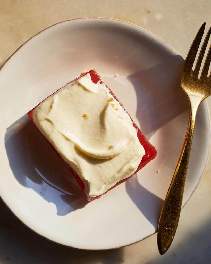 A watermelon strawberry jello bar with layers of pretzel shortbread and whipped cream sits on a dessert plate