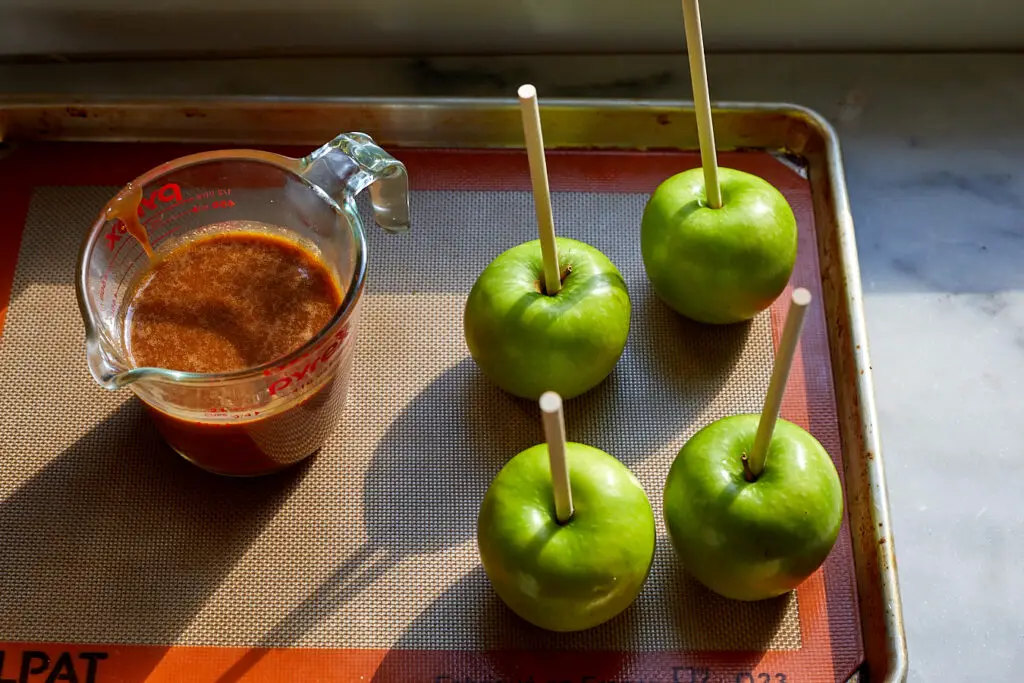 green granny smith apples sitting on a baking sheet next to a measuring cup of caramel