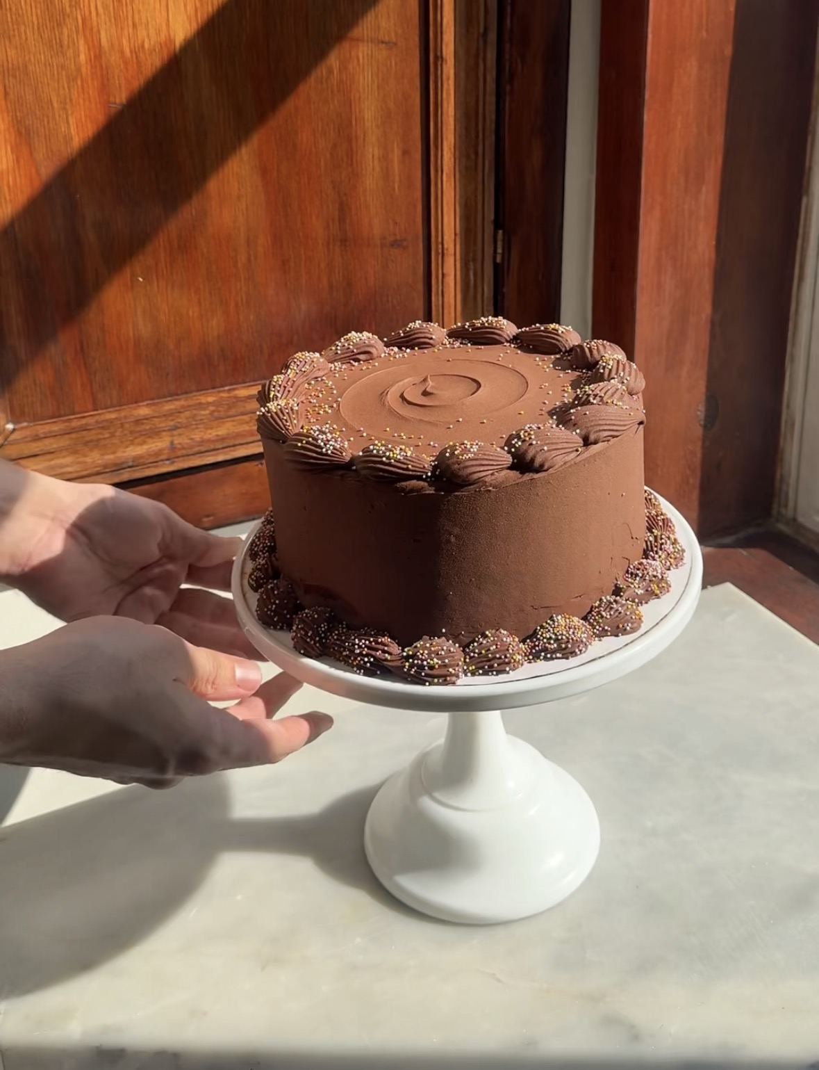 Hands placing a chocolate birthday cake atop a white cake stand onto a marble surface