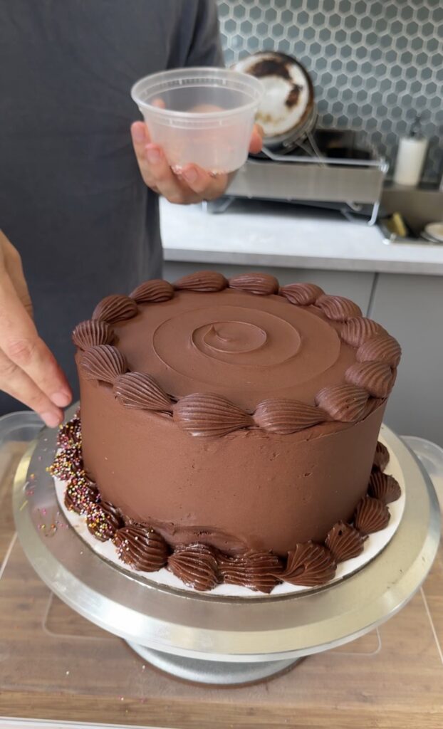 A hand sprinkles rainbow nonpareils on the buttercream piping of a chocolate birthday cake